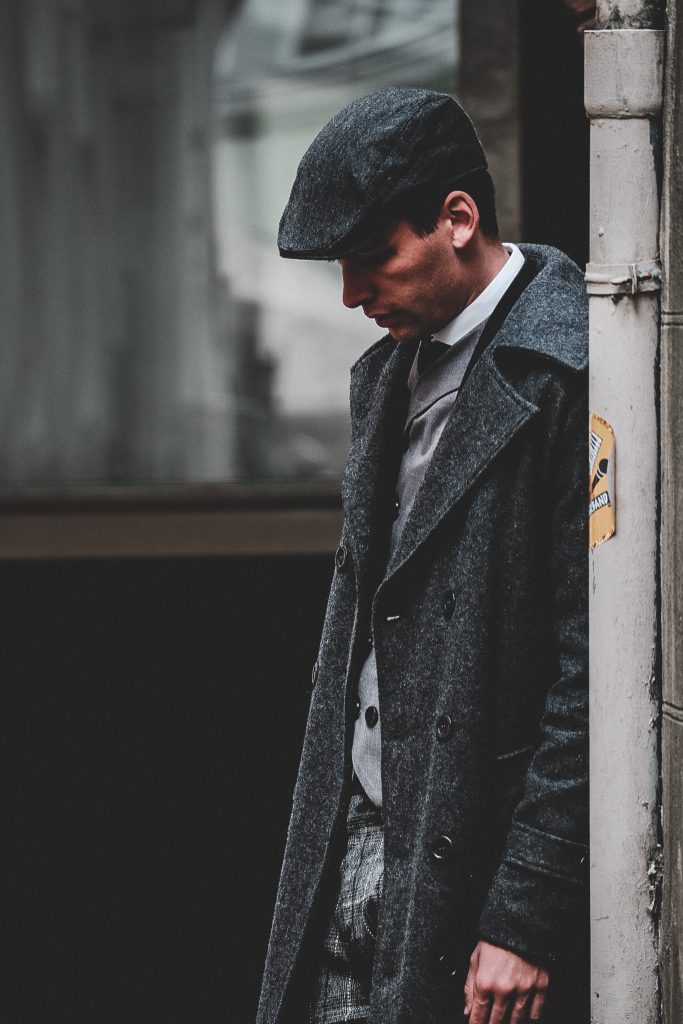 A man wearing a flat cap and a grey overcoat, clothing often seen at Cheltenham.