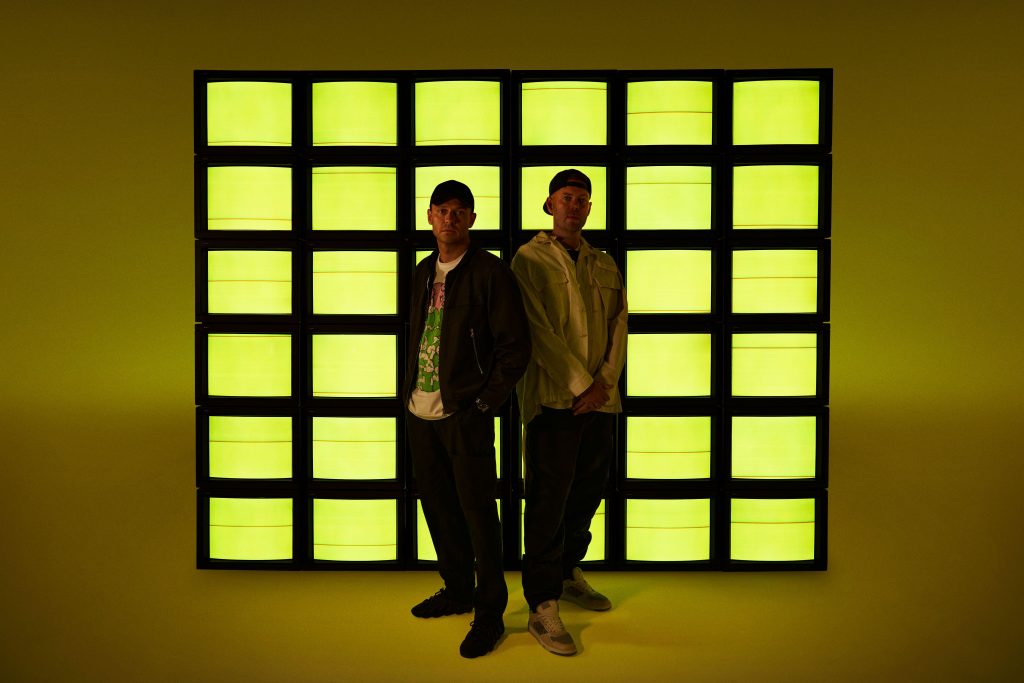 Solardo standing in front of a wall made of TV screens bathed in yellow.