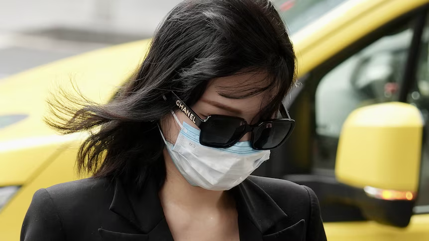 A young woman in a dark coat and white face mask walking past a yellow taxi, head lowered