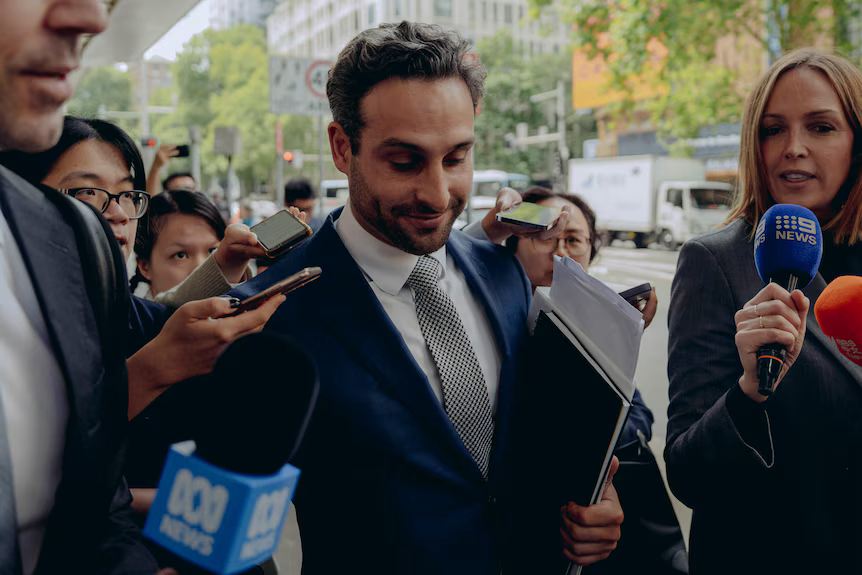 A man in a suit reading a document while reporters with microphones crowd around him outside a courthouse
