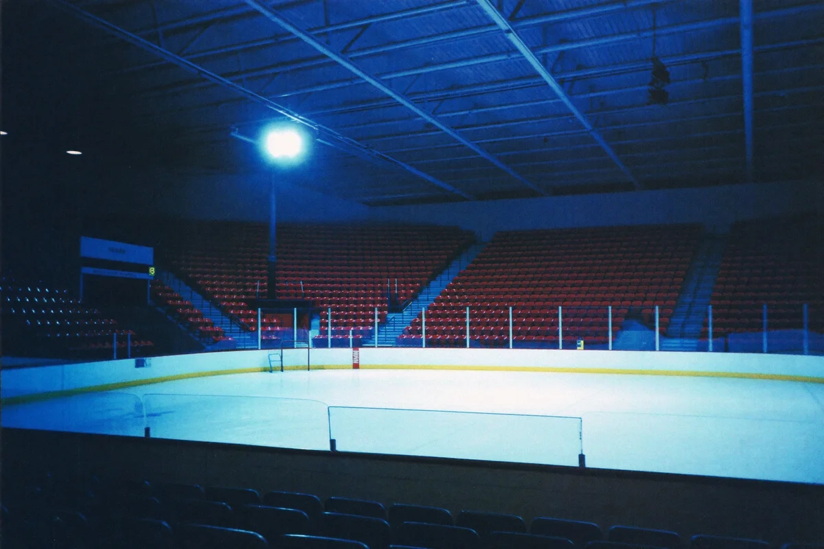 An empty vintage ice-hockey arena bathed in deep blue light, with white ice and rows of red seats in shadow
