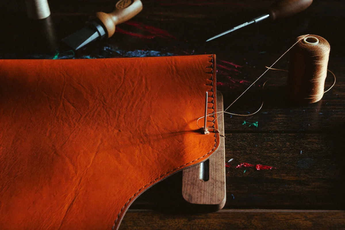 Close-up of orange leather being saddle-stitched with a needle and waxed thread on a wooden stitching clamp, with a spool of thread nearby