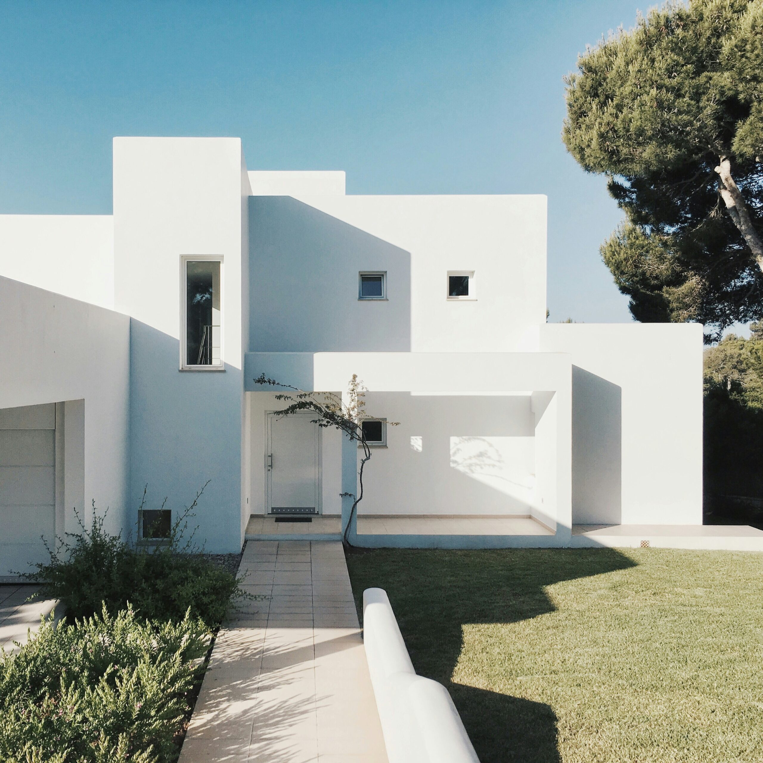 Modern white building with rectangular forms, a green lawn, sparse shrubbery, and a large tree under a clear sky.