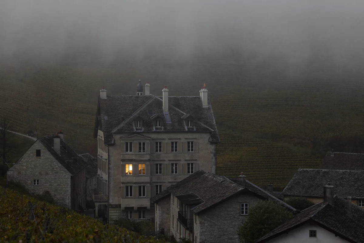Stone village in thick fog with a large shuttered manor house, a single upstairs window lit